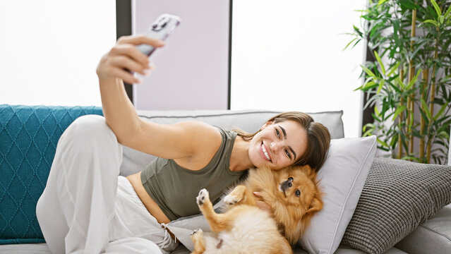 Joyful, young hispanic woman comfortably sitting on her living room sofa at home with a smile. enjoying indoor time taking a happy selfie picture with her pet dog using smartphone technology.