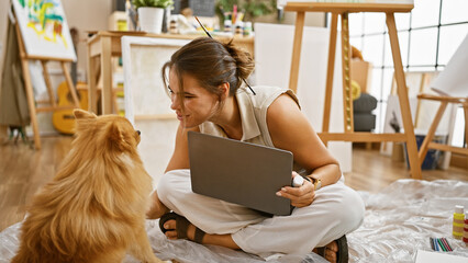 Smiling young hispanic woman artist, with her dog, sitting on studio floor, engrossed in painting...