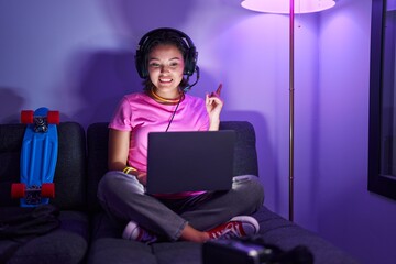 Hispanic young woman playing video games sitting on the sofa smiling happy pointing with hand and finger to the side