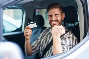 Hispanic man with beard driving car holding credit card screaming proud, celebrating victory and...