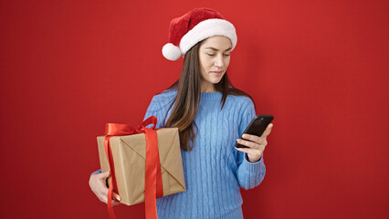 Young caucasian woman wearing christmas hat using smartphone holding gift over isolated red background