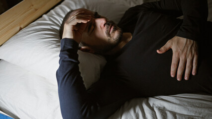 A bearded, bald hispanic man lying in bed with a pained expression on his face in a dimly lit room...