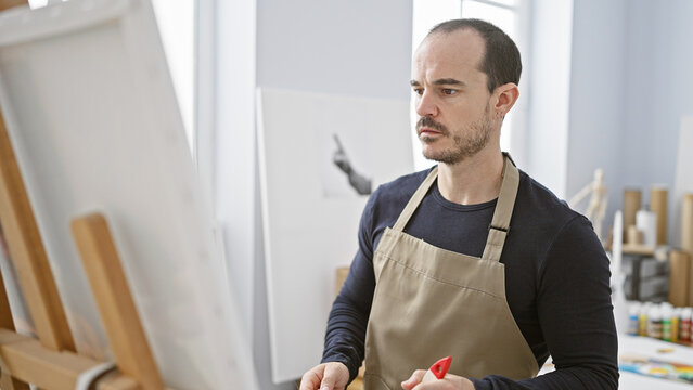 Focused man with beard in apron painting on canvas in art studio, embodying creativity.