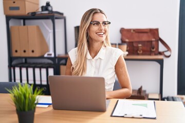 Young blonde woman working at the office wearing glasses looking away to side with smile on face,...