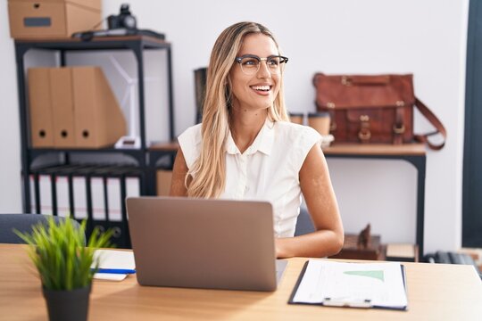 Young blonde woman working at the office wearing glasses looking away to side with smile on face, natural expression. laughing confident.