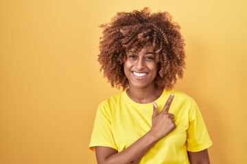 Young hispanic woman with curly hair standing over yellow background cheerful with a smile of face pointing with hand and finger up to the side with happy and natural expression on face