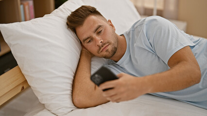 A young hispanic man with a beard lounging in a bedroom, looking at a smartphone while reclining on...