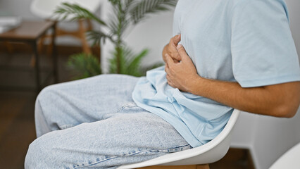 Hispanic man with stomach pain sitting in a modern indoor waiting room.