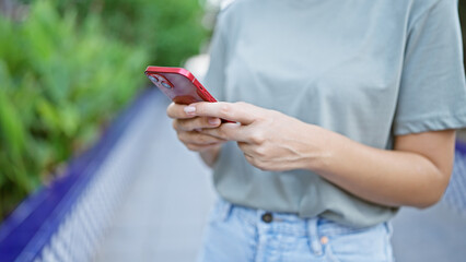 A young woman uses a smartphone while leisurely standing outdoors surrounded by greenery.