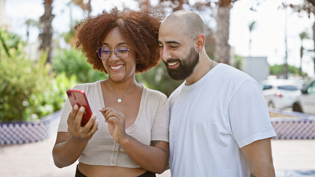 Confident, happy interracial couple smiling together, standing in sunlit park with their smartphone, effortlessly sharing love and laughter outdoors