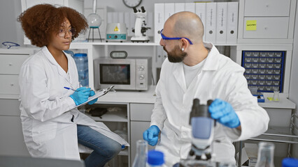 Inside the buzzing lab, two committed scientists, man and woman, analysis partners, seated together, gloved hands busy writing on clipboard, eyes glued to the microscope in serious research