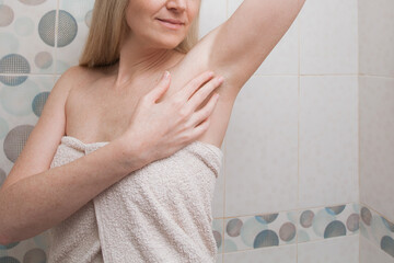 Naklejka premium Positive beautiful woman with blond hair and in a white towel, touches her shaved armpits in the bathroom. Image on the theme of health, hygiene and personal care.