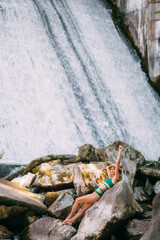 A slender sweet girl with blond hair, tanned skin, dressed in a green stylish swimsuit and with a hat, sits on stones against the backdrop of a picturesque waterfall and rests in the summer.