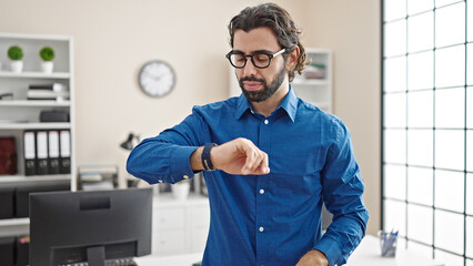Young hispanic man business worker looking watch with serious face at the office