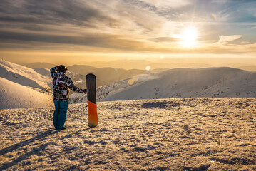Snowboarder with board standing on the slope in winter mountains and watching beautiful sunset and...