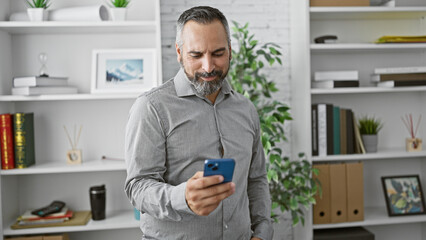 Mature hispanic man with beard and grey hair using smartphone in modern office interior.