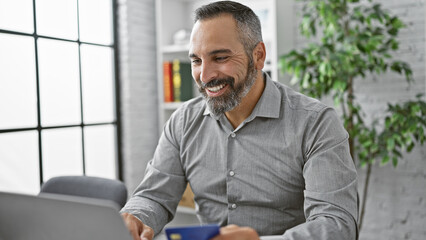 Smiling mature hispanic man with gray beard working on laptop in modern office, holding credit card.