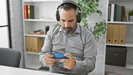 Handsome senior hispanic man with a beard and grey hair wearing headphones, using a smartphone in an office setting.