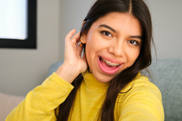 Happy Latin female teenager with braces taking a selfie at home.