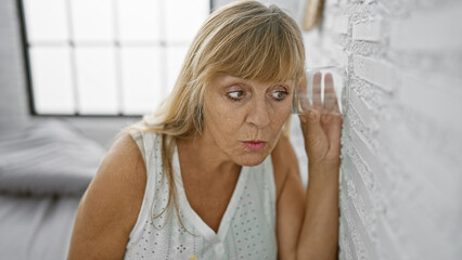 Curious middle age blonde woman eavesdropping, spying through bedroom wall with glass, indoor nosy...