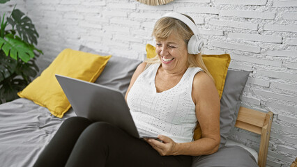 Blissful middle age blonde woman relaxing in her cozy bedroom, sitting on bed with laptop, enjoying music through headphones, radiating happiness and confidence