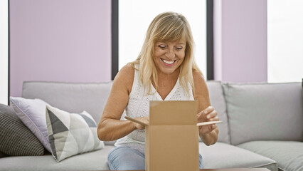 Joyful middle age blonde woman delightfully unpacking a cardboard box at her beautiful home,...