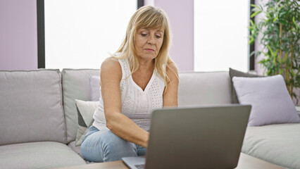 Obraz premium In her cozy home, a serious and focused middle age blonde woman is sitting on the sofa, totally absorbed, using her laptop in the living room's welcoming interior.