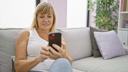 Beautiful middle-aged blonde woman sitting on a sofa at home, enjoying the joy of texting on her smartphone, a cheerful expression of happiness on her face