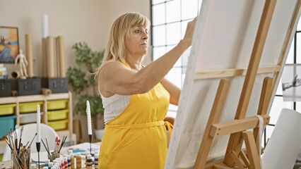 Beautiful middle age blonde woman artist seriously concentrating on drawing in her indoor art studio