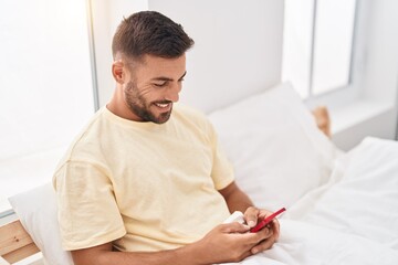 Young hispanic man using smartphone sitting on bed at bedroom