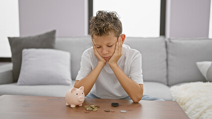 Adorable blond boy, sitting solemn and sad at home, sceptically studying coins and piggy bank,...