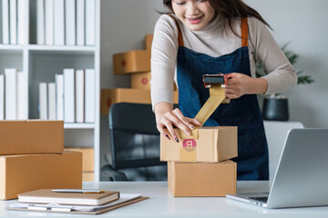 Young asian woman taping up a cardboard box in home office SME e-commerce business, relocation and...