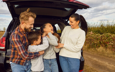 Portrait of happy smiling family of four with two kids boy and girl having fun and hugging standing near the trunk of their car enjoying weekend in nature. Vacation and road trip concept