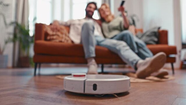 An adult couple with a remote control having fun sitting on a sofa by the window in the living room at home, in the foreground a robot vacuum cleaner cleans the house.