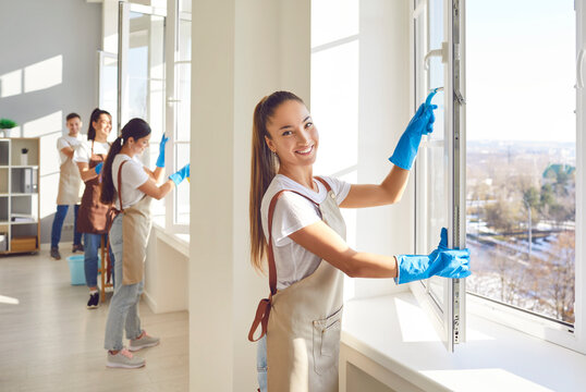 Portrait of a young cleaner girl from professional cleaning service looking cheerful at camera and smiling while cleaning windows in the house with a team of janitors in background.