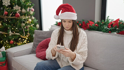 Obraz na płótnie Canvas Young beautiful hispanic woman using smartphone sitting on sofa by christmas tree at home