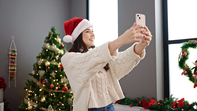 Young beautiful hispanic woman make selfie by smartphone standing by christmas tree at home