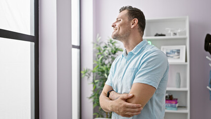 Fototapeta na wymiar Handsome man with arms crossed standing in a contemporary home interior, looking through window.