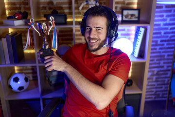 Young hispanic man playing video games holding trophy looking positive and happy standing and...