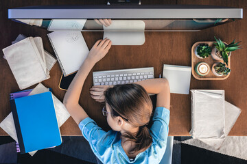 A tired girl lies on a table in front of a computer with notepads and books.