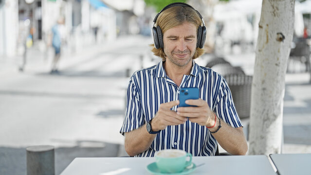A middle-aged man with blond hair listens to music on headphones, using a smartphone at a city street cafe.