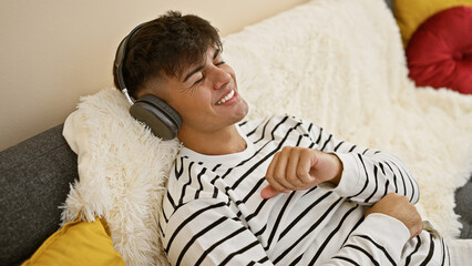 Young hispanic man sitting on sofa listening to music dancing at home