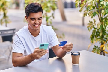 Young hispanic man using smartphone and credit card sitting on table at coffee shop terrace