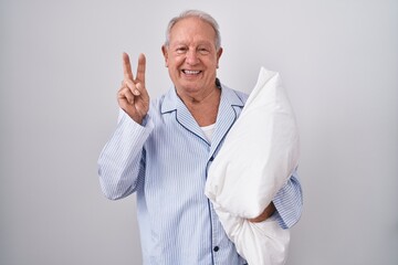 Senior man with grey hair wearing pijama hugging pillow smiling with happy face winking at the...