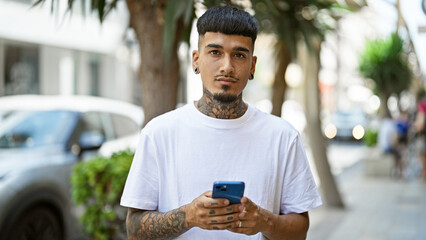 Cool and tattooed young latin man concentrated on his smartphone, texting while standing outdoors on a sunny city street sporting serious expression and relaxed lifestyle