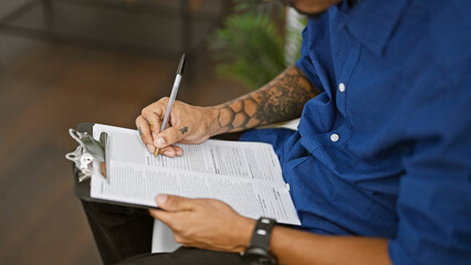 Concentrated hispanic man with tattooed hands sitting in chair, seriously writing on clipboard in...