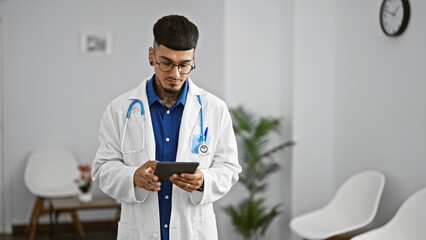 Relaxed, young latin man doctor tattoos reveals, working at his clinic, concentrated on internet...