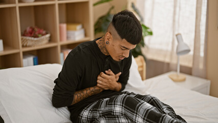 Handsome young latin man, tattooed and suffering, sitting on his bed gripping chest in agony from heart attack at home