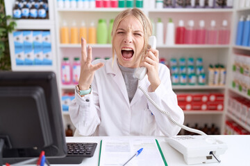 Young caucasian woman working at pharmacy drugstore speaking on the telephone crazy and mad...