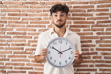 Arab man with beard holding big clock puffing cheeks with funny face. mouth inflated with air, catching air. - obrazy, fototapety, plakaty